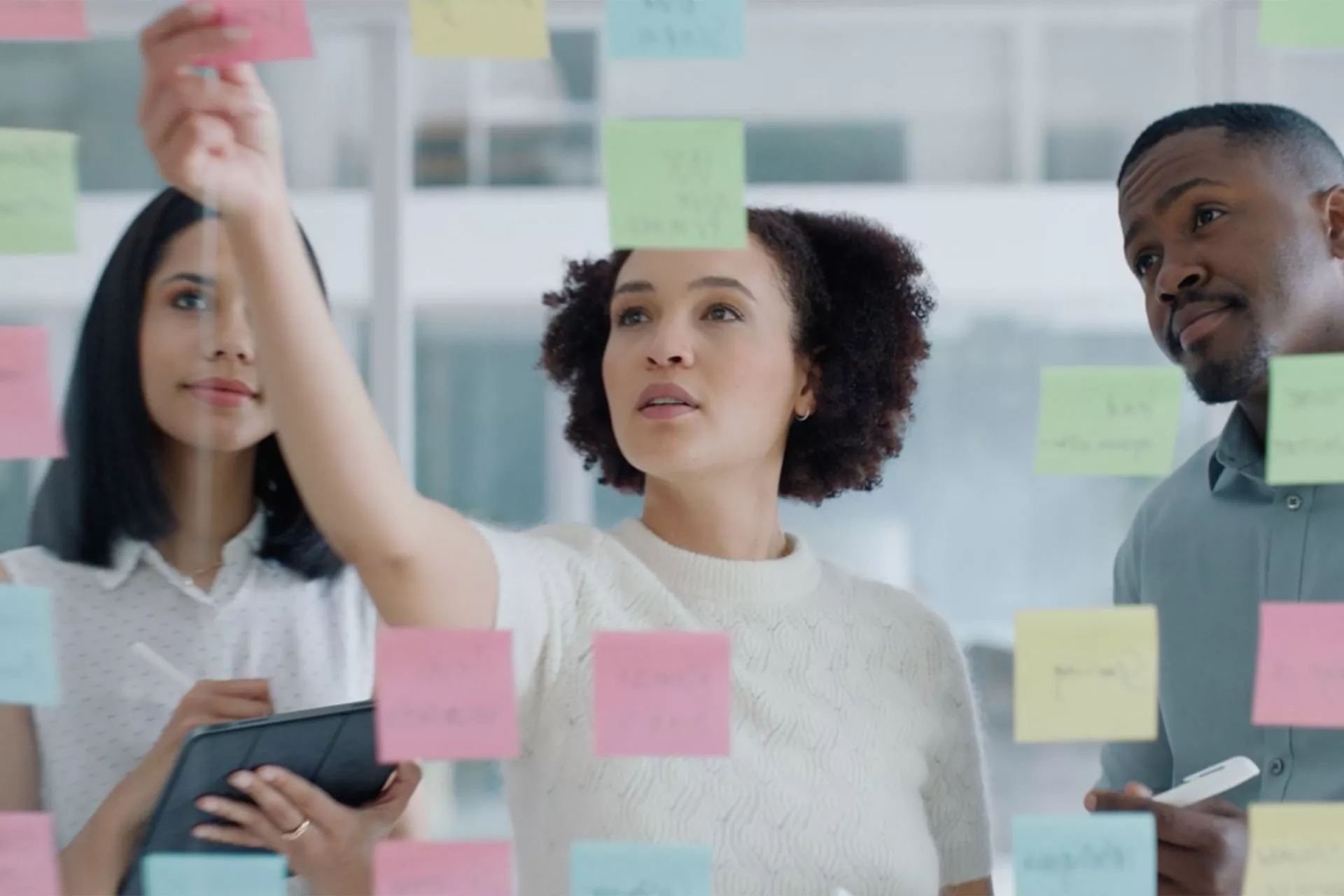 Three diverse professionals collaboratively brainstorming with sticky notes on a glass wall in a bright, modern office. A woman is actively placing a note while her colleagues observe and contribute to the discussion.
